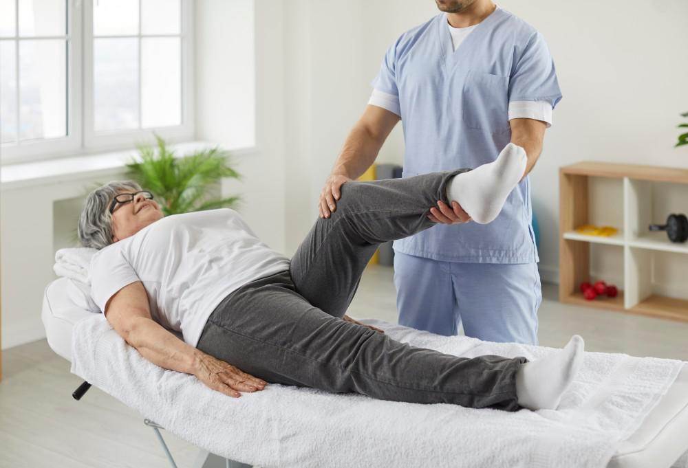 Physical therapist aiding a senior woman in leg exercises for her pain management plan in a clinic.